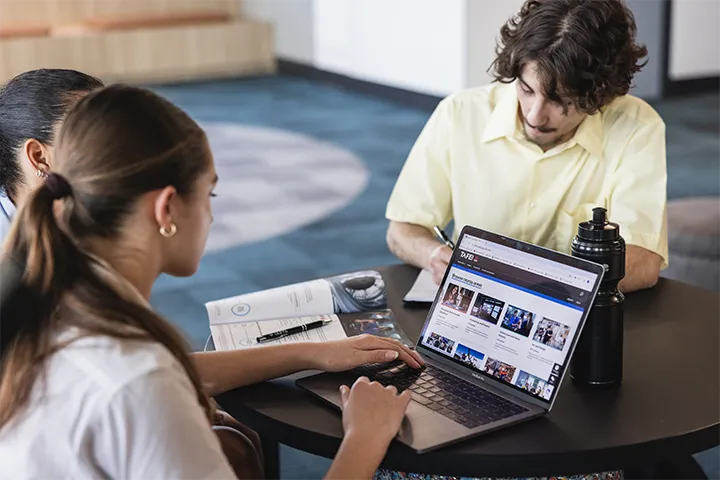 School students sit together at a round table viewing the TAFE NSW website on a laptop while discussing course options, with notebooks, pens, and study materials spread across the table in a modern learning space.