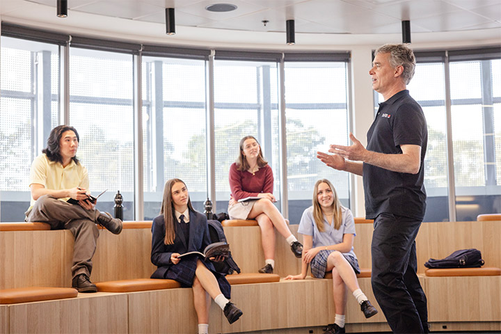 Teacher speaks to a group of school students seated on curved wooden tiered benches in the TAFE NSW amphitheatre space, with large windows providing natural light and personal belongings placed beside the students.