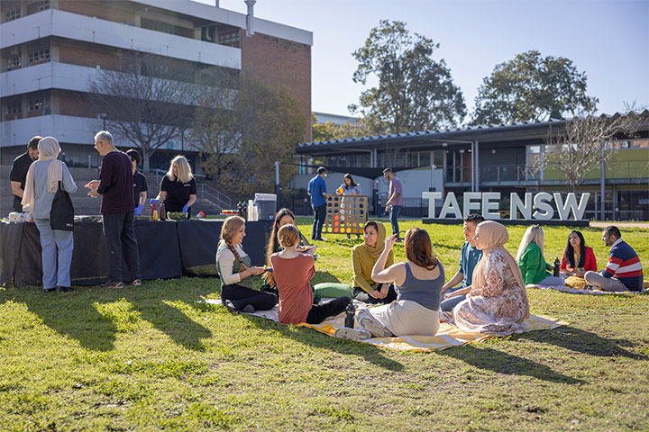 People sit on picnic blankets and gather around serving tables on a grassy area during a ɫֱBBQ event, with campus buildings and a large ɫֱsign visible in the background under bright afternoon sunlight.