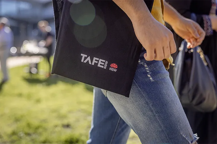 Close‑up of a black TAFE NSW tote bag hanging from a person's shoulder, with the TAFE NSW logo printed in white and red, captured outdoors on a sunny day with grass and people in the background.