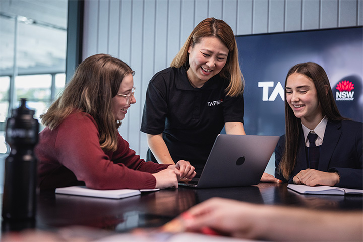 Advisor stands at a table guiding two students during a meeting, with a laptop open in front of them, notebooks on the table, and a TAFE NSW screen displayed in the background of the modern meeting room.