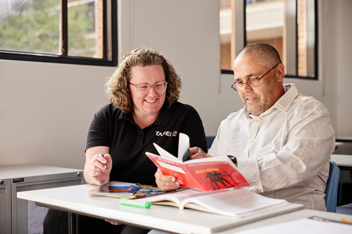 TAFE NSW adult learner sits at a desk reading from an open book while a teacher provides one‑on‑one guidance, with additional learning materials and notes spread across the table in a bright classroom setting focused on improving literacy skills.