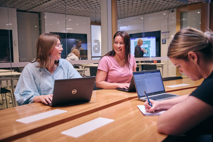 Students sit at a long wooden table inside a modern Connected Learning Centre, working on laptops and taking handwritten notes while collaborating and talking together, with glass walls, digital screens, and other learners visible in the background of the contemporary study space.