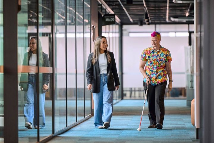 Two people walk side by side through a modern campus corridor with glass walls, one using a white mobility cane, highlighting an accessible and inclusive learning environment with clean lines, reflective surfaces, and natural light.
