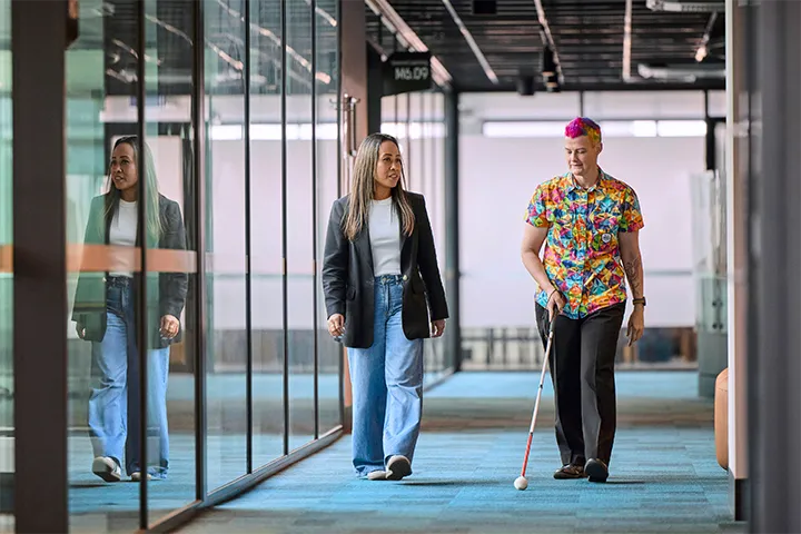 Two people walk side by side through a modern campus corridor with glass walls, one using a white mobility cane, highlighting an accessible and inclusive learning environment with clean lines, reflective surfaces, and natural light.
