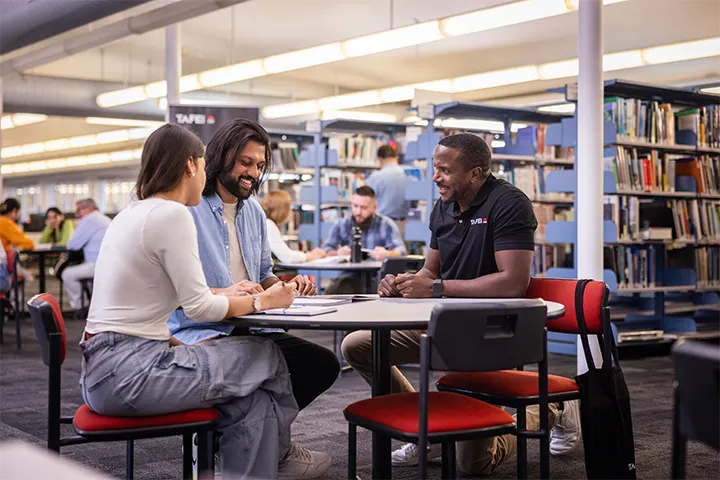 TAFE NSW staff member providing library learning support to two students at a circular table, with bookshelves and other learners in the background.