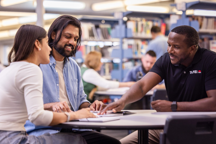 TAFE NSW staff member providing library learning support to two students at a circular table, with bookshelves and other learners in the background.