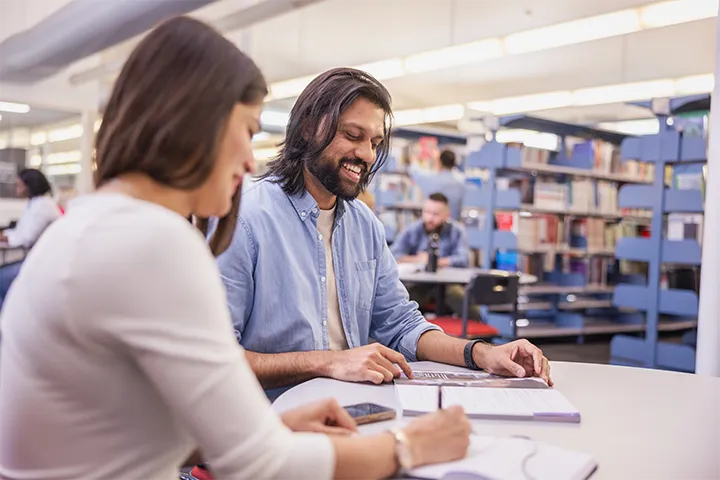 Two individuals seated at a table in a library, engaged in discussion, with bookshelves and other students studying in the background.