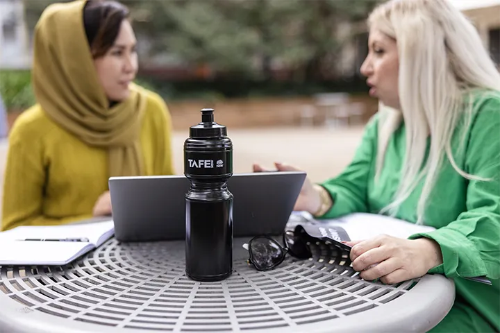 Two students sitting at a circular outdoor table on campus with laptops, a TAFE NSW branded water bottle, notebooks, and sunglasses.