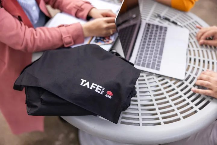 Close-up of a black TAFE NSW branded t-shirt with white and red logo on an outdoor table, with two students studying using laptops and notebooks.