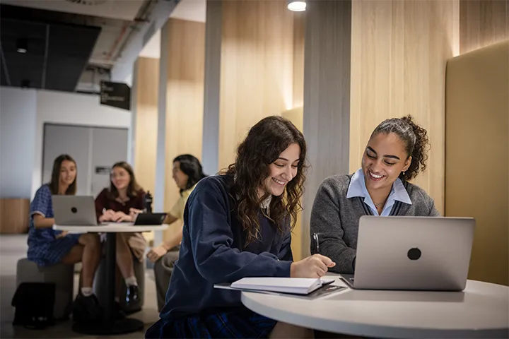Students working in study pods within a modern indoor learning space, seated at round tables with laptops and notebooks, surrounded by cushioned seating and wooden wall paneling.