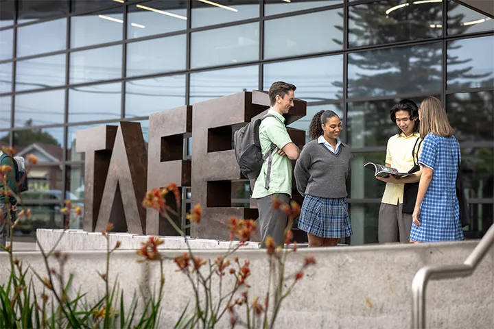 Group of four students in school uniforms talking in front of a large 'TAFE NSW' sculpture on campus, with one student holding an open book, modern glass building and red flowering plants in the background.