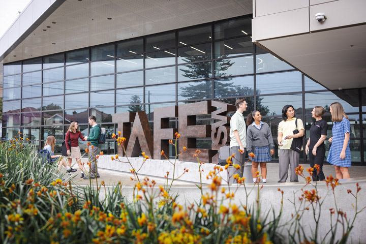 Students stand and talk outside a modern TAFE NSW campus building beside large sculptural “TAFE” letters, with glass façades, landscaped gardens, and pathways in the foreground creating a welcoming campus entrance scene in daylight.