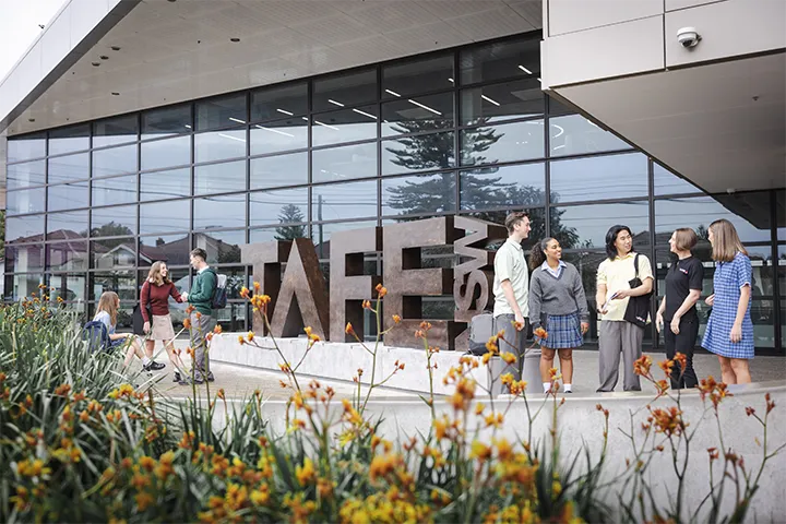 Students stand and talk outside a modern TAFE NSW campus building beside large sculptural “TAFE” letters, with glass façades, landscaped gardens, and pathways in the foreground creating a welcoming campus entrance scene in daylight.