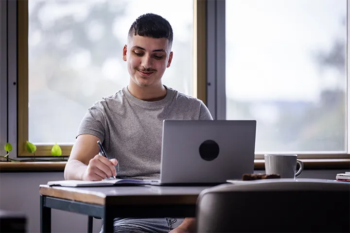 Person working from home at a desk with a laptop and notebook, writing with a pen, surrounded by natural light from large windows and indoor plants, with a coffee mug and food items on the desk.