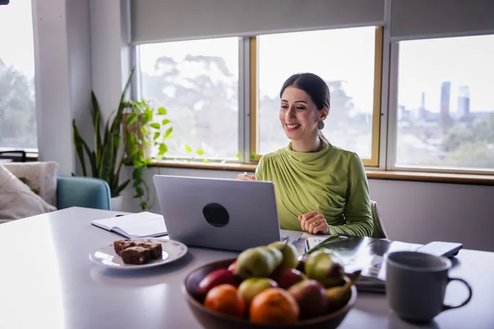 Person working from home at a desk with a laptop, notebook, coffee mug, and plates of fruit and cake, seated near large windows with natural light and indoor plants.
