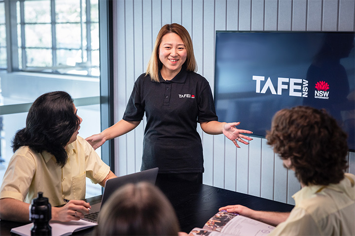Teacher in a TAFE NSW classroom stands at the front of the room speaking to two future students seated at a table with notebooks and a laptop, with a TAFE NSW screen displayed behind her in a modern learning space.