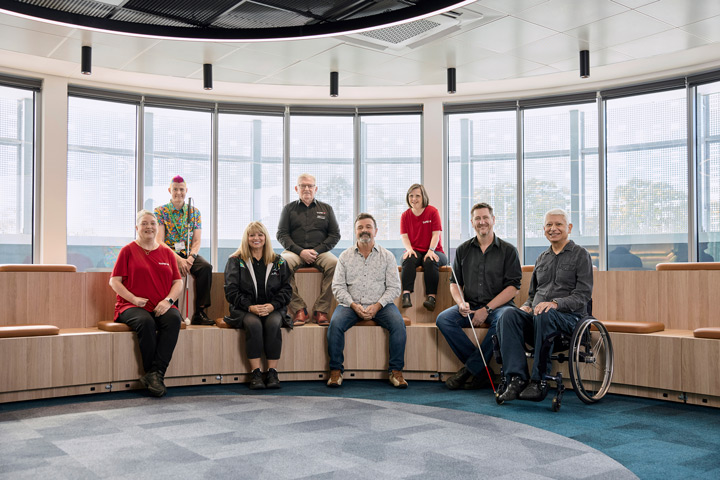 Eight individuals are sitting together. One man is in a wheelchair, one woman and man have a guide cane. They are all smiling and looking at the camera.