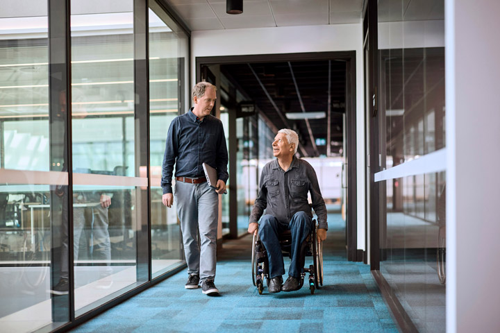 Two individuals in a modern ɫֱoffice hallway, one walking with a laptop and the other using a wheelchair, both dressed in casual business attire, highlighting diversity and inclusion in an accessible workplace.