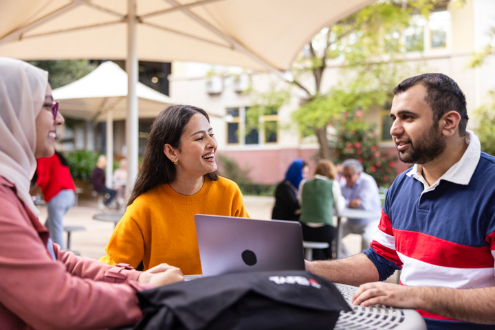 Three students sitting under a large umbrella at a circular outdoor table, discussing with a laptop open, surrounded by greenery and campus buildings.