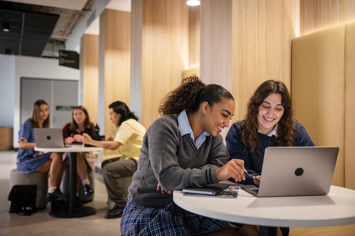 Students using study pods in a modern indoor learning space with round tables, laptops, cushioned seating, and wooden wall paneling, engaged in collaborative study and discussion.