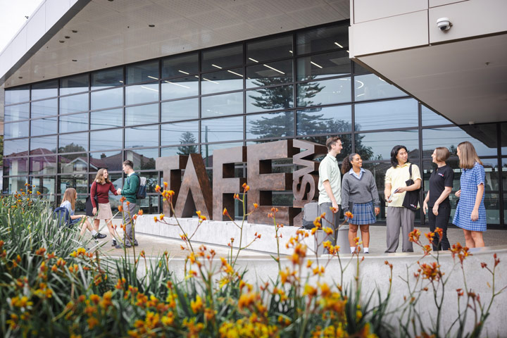Group of students and a teacher standing in front of a large 'TAFE NSW' sculpture on campus, with modern glass buildings and flowering plants in the background.