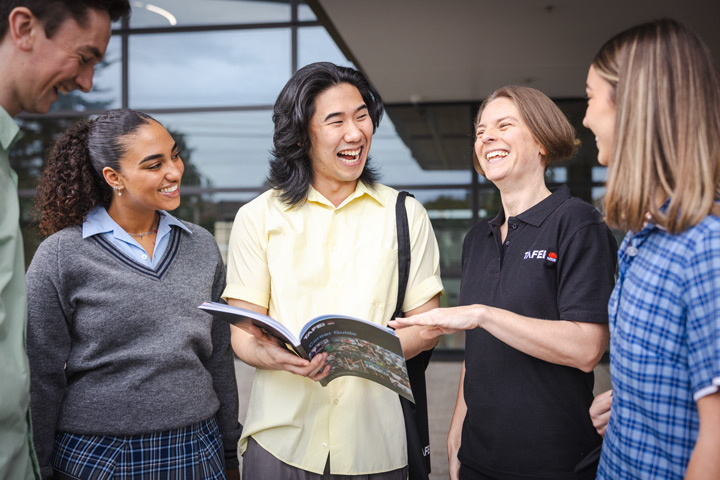 Group of students standing outside a modern building with large glass windows, listening to a person in a ɫֱshirt who is pointing at an open book.