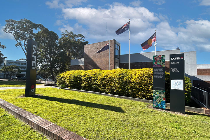TAFE NSW Belmont campus featuring a grassy area, two vertical signs—one reading “TAFE NSW” and the other “TAFE Belmont Pacific Highway” with artwork and an info panel. A building, flagpoles, and trees are visible in the background under a partly cloudy sky.