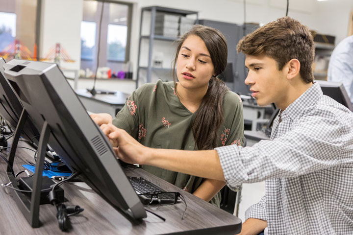 Two students sit at a desk together. They’re working on a laptop and are engaged in their task.