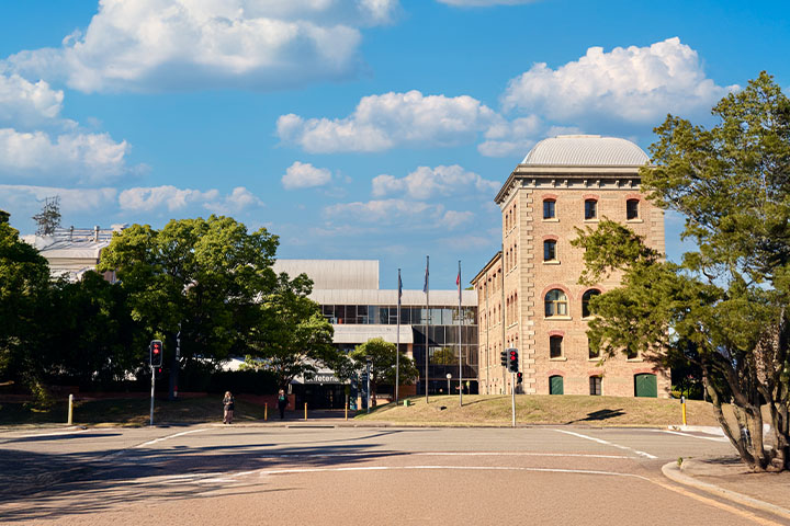 TAFE NSW Hamilton campus featuring a heritage-style brick building with arched windows and a domed roof alongside a modern glass-fronted structure, set near a tree-lined street with traffic lights under a bright blue sky with scattered clouds.