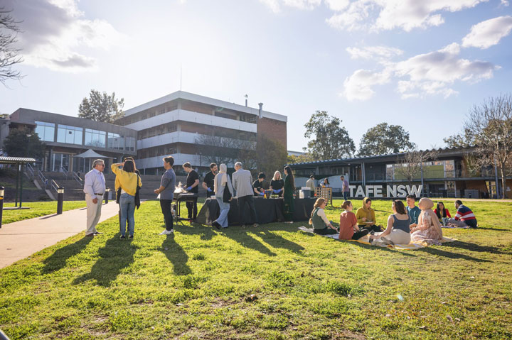 A group of people socialise outdoors on a sunny day near a TAFE NSW campus. There are surrounding buildings, a table with food, and small groups seated on the grass.
