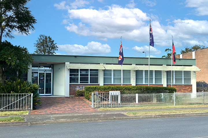 Exterior view of the TAFE NSW Wauchope campus showing a single‑storey building with green roofing, brick and rendered walls, large front windows, and three flagpoles displaying flags at the entrance, set behind a low fence with landscaped gardens and a footpath along the street.