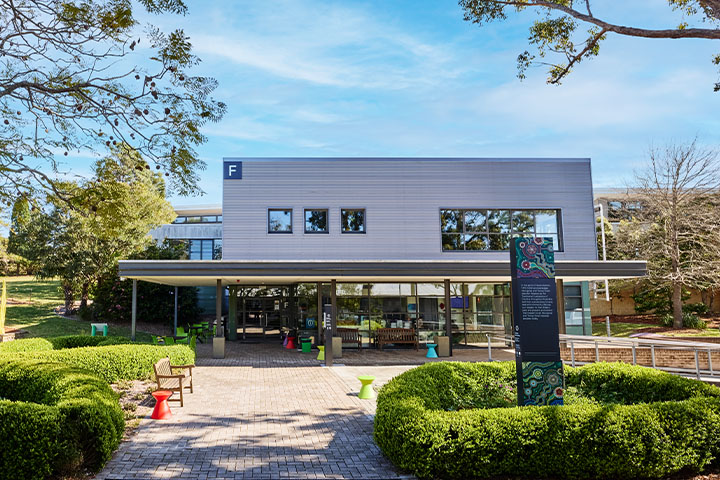 TAFE NSW Yallah Campus Building F with large windows, a covered entrance, outdoor seating, and landscaped greenery including trees and bushes, under a partly cloudy sky.
