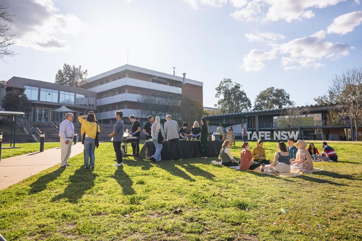 Group of people socialising outdoors on a sunny day near a TAFE NSW campus building, with tables set up and small groups seated on the grass.