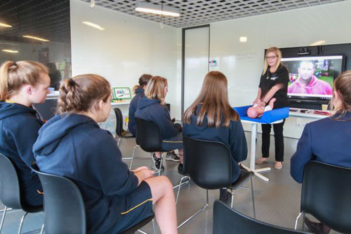 A group of students seated in chairs, attentively watching a teacher conduct a presentation in a classroom setting.