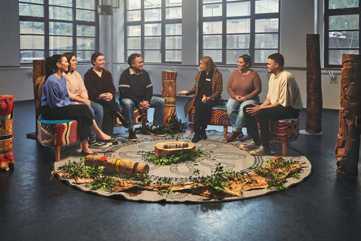 An Aboriginal mob sitting in a yarning semi circle, engaging in conversation. There is a round rug they are sitting around which is placed leaves, plants and other culturally significant pieces.