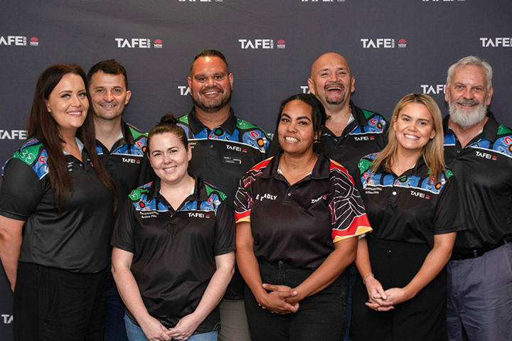 A group of eight Aboriginal TAFE NSW workers smiling to the camera and wearing TAFE NSW shirts with a range of colourful Indigenous designs.