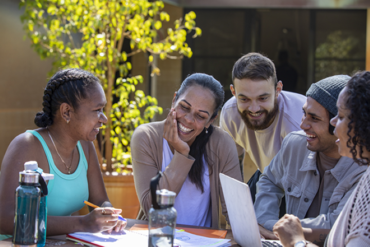 A group of smiling and laughing Aboriginal students sit outside at a table. There are green trees in the background and water bottles on the table.