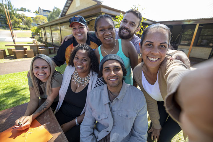 A group of 7 Aboriginal people are taking a selfie outside on a clear and sunny day.