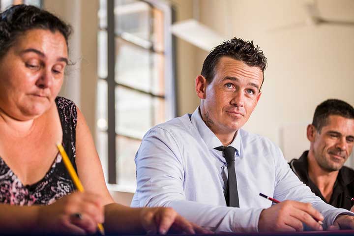 A panel of Aboriginal men and one women sit at a table with pencils in their hands.
