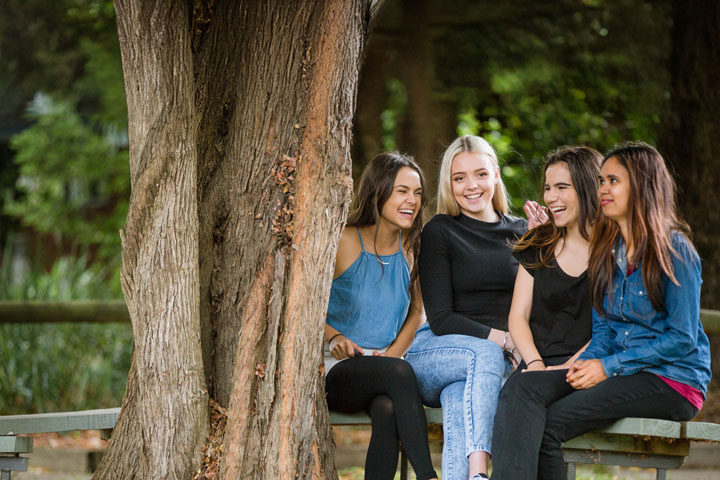 Four young women of Aboriginal heritage sitting on a bench outdoors, smiling and chatting together near a large tree. The scene is surrounded by greenery and fallen leaves.