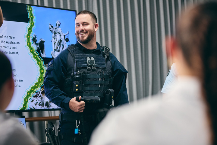 Person in tactical gear standing in front of a presentation screen with partially visible text.