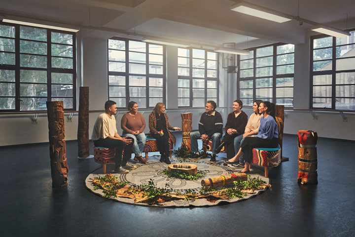 An Indigenous mob sitting in a yarning semi circle, engaging in conversation. There is a round rug they are sitting around which is placed leaves, plants and other culturally significant pieces.