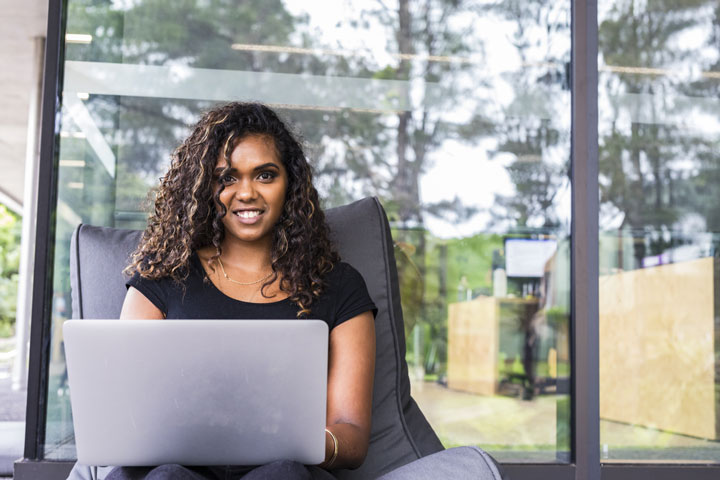An Aboriginal student with curly long hair sits on a chair with a laptop on her lap. Behind her are large glass windows showing trees and greenery outside.