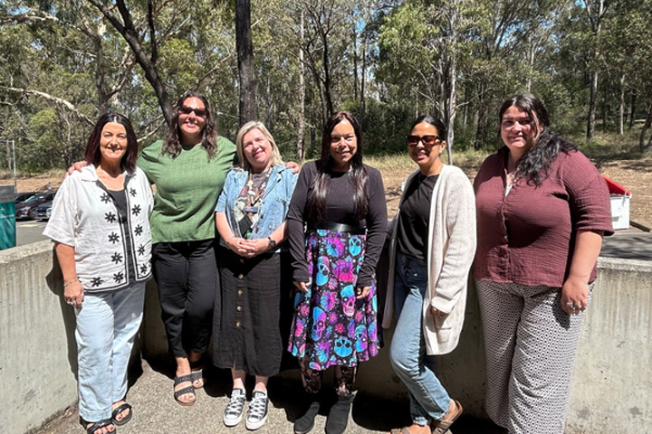 Eight Aboriginal and Torres Strait Islander women standing together outdoors in solidarity with trees in the background.