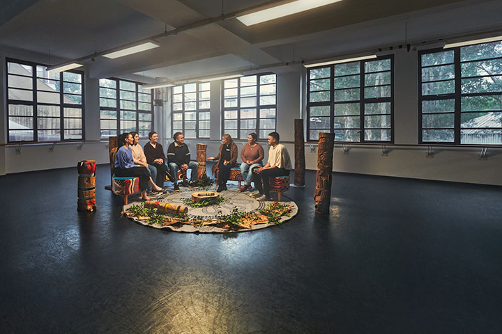 An Indigenous mob sitting in a yarning semi circle, engaging in conversation. There is a round rug that they are sitting around, leaves, plants and other culturally significant pieces.
