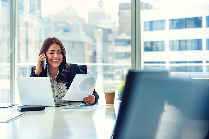 A smiling woman is working in a modern, well-lit office. She is talking on the phone and looking at a document.