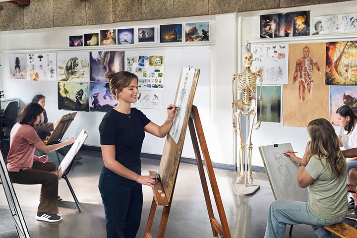 A group of students are in an art workshop sketching a skeleton model that is propped on a stand at the front of the class. There are lots of art works plastered on the walls around them.