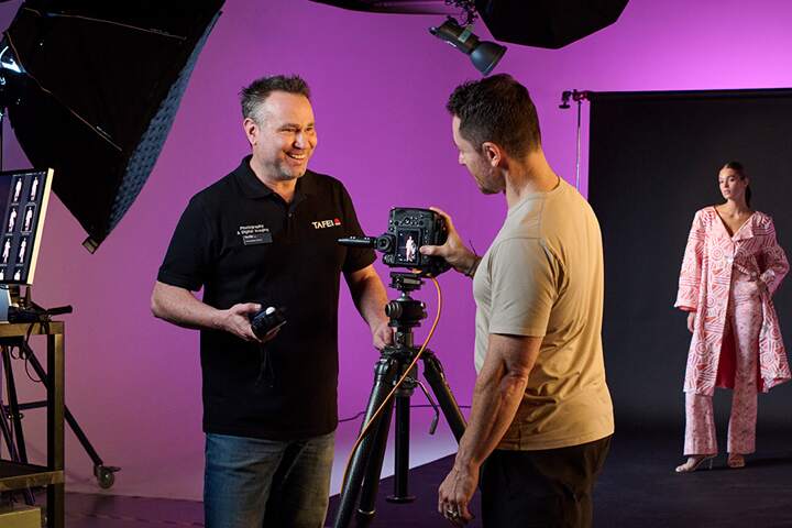A photography teacher and student working together in a studio setting, with a model posing in the background.
