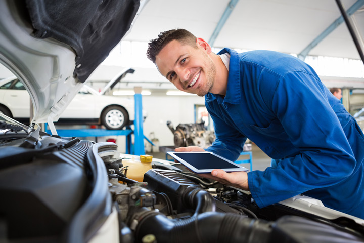 A male student in a garage is dressed in blue overalls. He holds a tablet and observes a car with a positive expression.